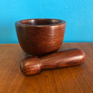 May include: A wooden mortar and pestle set. The mortar is a deep bowl shape, and the pestle is cylindrical with a rounded end. Both pieces are made of a dark, reddish-brown wood with visible grain. The set is designed for grinding and mixing ingredients.