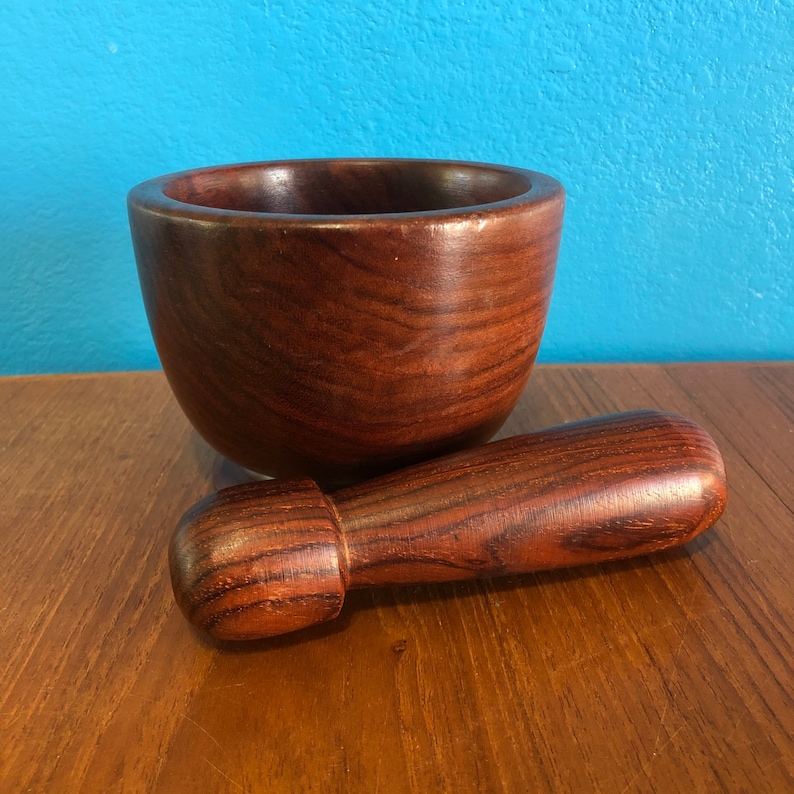 May include: A wooden mortar and pestle set. The mortar is a deep bowl shape, and the pestle is cylindrical with a rounded end. Both pieces are made of a dark, reddish-brown wood with visible grain. The set is designed for grinding and mixing ingredients.