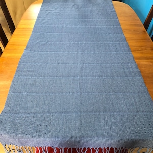 May include: A blue woven table runner with a geometric pattern and fringe. The runner is made of a soft, textured fabric and features a diamond pattern at the ends and horizontal stripes across the center. The runner is displayed on a wooden table.
