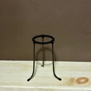 Puede incluir: Un soporte de trípode de metal negro con una parte superior circular y tres patas curvas. El soporte está diseñado para sostener un recipiente y es adecuado para uso en laboratorio o decorativo.