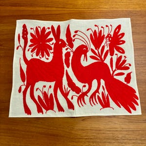 May include: A white linen textile featuring red embroidered designs of a rabbit, a bird, flowers, and a butterfly. The embroidery is detailed and the design is symmetrical. The textile is likely a decorative piece or a kitchen towel.