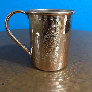 Peut inclure: Une tasse en cuivre martelé avec un motif de hibou. La tasse a une poignée et est faite d'un métal brillant.