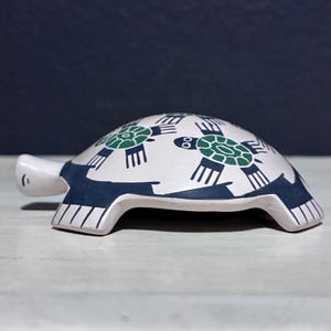 May include: A ceramic turtle figurine with a white body, dark blue accents, and green turtle designs. The turtle has a rounded shell and a small head. The figurine is on a white surface with a dark blue background.