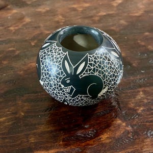May include: A black, round, carved stone vessel with a rabbit design. The vessel has a circular opening at the top and is decorated with a white, web-like pattern. The rabbit is carved in a dark color, and the overall design is intricate and detailed.