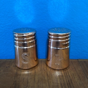 May include: Two copper-coloured salt and pepper shakers. They have a cylindrical shape with a ridged design and a perforated top. The salt shaker is marked with an "S", and the pepper shaker is marked with a "P".