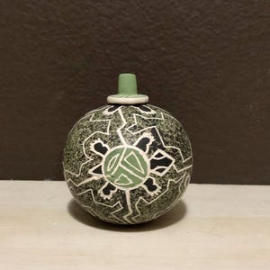 May include: A round, ceramic decorative jar with a green and black geometric design. The jar has a small, green stopper. The design includes a central green shape surrounded by black and white patterns. The jar is likely handmade and has a unique, artistic appearance.