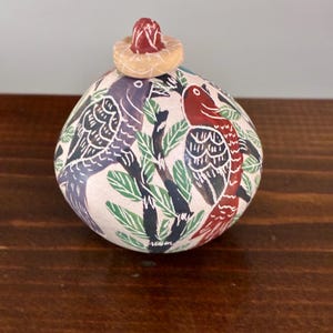 Op de afbeelding: Een ronde, handbeschilderde keramische pot met deksel en een lichtroze basiskleur. Het oppervlak is versierd met donkerpaarse en rode vogels op donkere takken tussen groene bladeren. Het deksel heeft een kleine, roodkleurige knop.