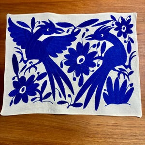 May include: A rectangular white textile featuring a vibrant blue embroidered design. The design includes two stylized birds, flowers, and foliage. The embroidery is detailed and creates a striking contrast against the white background.