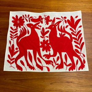 May include: White square textile with a vibrant red embroidered design. The design features two stylized animal figures facing each other, surrounded by floral and foliate elements. The embroidery is detailed and the overall aesthetic is folk art.
