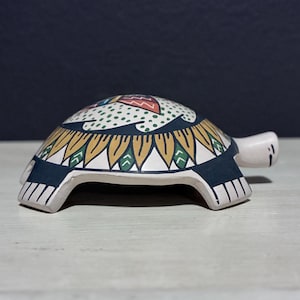 May include: A ceramic turtle figurine with a painted design. The turtle's shell features a pattern of yellow, blue, and green geometric shapes. The turtle's head and feet are a light beige color. The figurine is on a white surface.