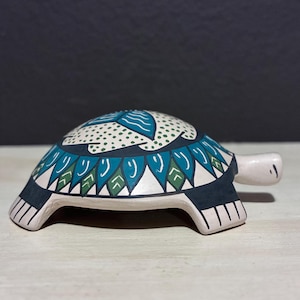 May include: A ceramic turtle figurine with a white body and a patterned shell. The shell features a design in teal, green, and black, with a dotted white background. The turtle has a small head and four stubby legs.