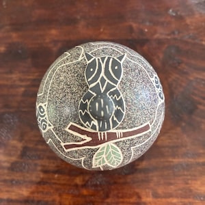 May include: A round, dark grey stone paperweight featuring a carved owl perched on a brown branch with light green leaves. The surface has a speckled texture with white etched lines outlining the owl, branch, and leaves in a circular pattern.