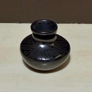 May include: A small, black ceramic vase with a rounded body and a narrow neck. The vase has a geometric pattern around the body. The vase is displayed on a light-coloured surface.