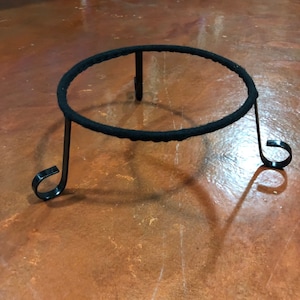 May include: A black metal plant stand with a circular top and three curved legs. The top is wrapped in a black, textured material. The stand is designed to hold a potted plant or decorative item.