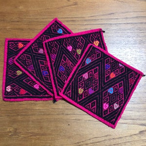 May include: Four pink and black square coasters with colorful geometric designs. The coasters are made of woven fabric and have a fringed edge.