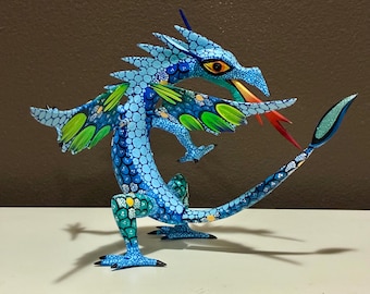 May include: A handcrafted, blue dragon sculpture with intricate details. The dragon features a textured body, green and yellow accents on its wings, and a red tongue. The sculpture is posed with its wings spread, and its tail curved.