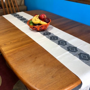 Peut inclure: Un chemin de table blanc avec un motif géométrique noir et gris. Le chemin de table est sur une table en bois avec un bol de fruits au centre.