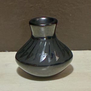 May include: A small, dark grey vase with a rounded body and a narrow neck. The vase has a series of vertical, carved lines that create a textured pattern around the body. The vase is sitting on a light-coloured surface.