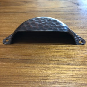 May include: A black hammered metal cabinet pull with a rounded top and a slightly curved shape.