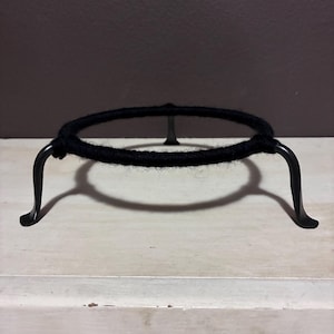 May include: A black metal trivet with a circular top wrapped in black yarn. The trivet has three curved legs and is designed to hold hot dishes. The trivet sits on a light-colored surface.