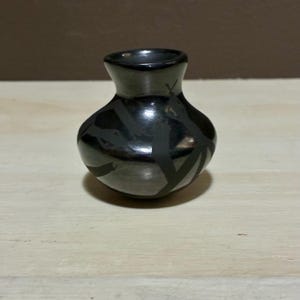 May include: A small, black ceramic vase with a rounded body and a narrow neck. The vase features a geometric pattern of dark grey lines. The vase is likely handmade and suitable for small floral arrangements or decorative purposes.