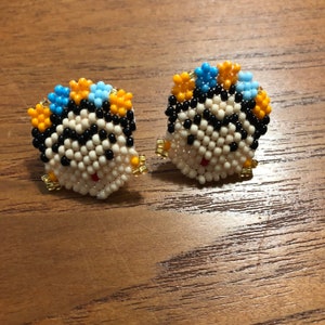 May include: A pair of beaded earrings with a cartoon face design. The earrings are made with white, black, yellow, and blue beads.