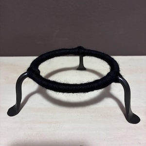 May include: A black metal stand with a circular top wrapped in black yarn. The stand has three curved legs and is designed to hold a round object. The stand is on a light-colored surface.