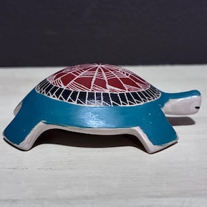 May include: A ceramic turtle figurine with a teal body and a red and white patterned shell. The turtle has a small head and tail, and is resting on a white surface.