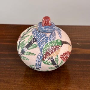 Op de afbeelding: Een ronde, lichtroze keramische pot met een deksel voorzien van een kleine rode en blauwe knop. Het oppervlak is versierd met een handgeschilderde afbeelding van een groene vogel met een lange snavel tussen donkergroene bladeren en blauwe takken.