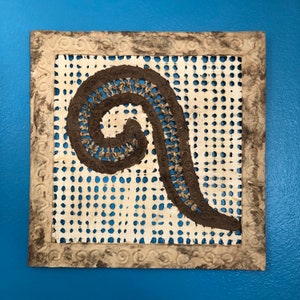 May include: A brown paper mache swirl design on a white background with a brown frame. The swirl is made of paper strips and has a textured look.