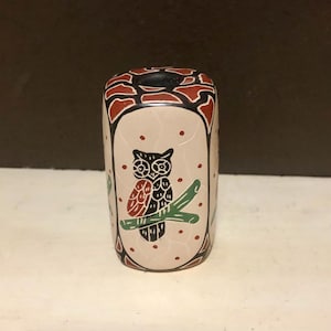 May include: A cylindrical ceramic owl figurine. The owl is perched on a green branch, with a red and black design. The background is a light beige color with red dots. The top of the figurine has a black opening.
