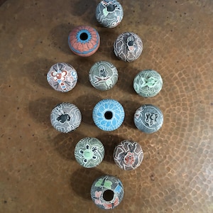 May include: A collection of decorative, hand-carved stone beads. Each bead features unique designs, including floral patterns, insects, and geometric shapes. The color palette includes shades of grey, blue, red, and brown. The beads are arranged on a copper-coloured surface.
