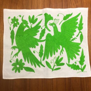 May include: White rectangular textile with vibrant green embroidered birds and floral designs. The birds are depicted with outstretched wings and detailed plumage. The embroidery is dense and creates a textured effect, showcasing a folk art style.