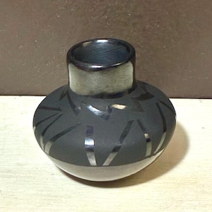 May include: A small, round, black ceramic vase with a wide opening and a short, cylindrical neck. The vase features a geometric design of silver-coloured lines. The neck is a lighter grey colour, and the vase sits on a white surface.