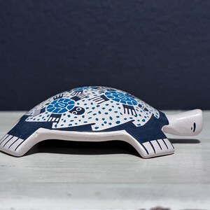 May include: A ceramic turtle figurine with a white base and a navy blue and white patterned shell. The shell features a repeating design of turtles and dots. The turtle's head and feet are white, set against a dark blue background.