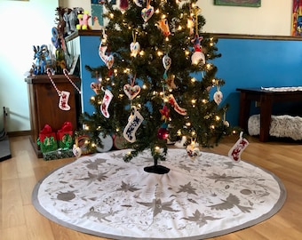 Pode incluir: Uma saia de árvore de Natal branca com um design floral bordado em cinza. A saia é colocada sob uma árvore de Natal decorada com luzes brancas e enfeites coloridos.