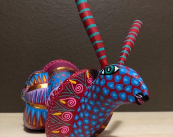 May include: A hand-painted, whimsical wooden sculpture of a creature with a red body, blue spots, and long red antennae with teal stripes. The sculpture has a detailed design with gold accents and a single green eye.