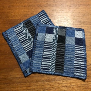 May include: Three blue and white woven coasters with geometric patterns. The coasters are made of a soft, textured fabric and have a fringed edge.