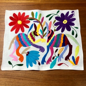 May include: A white linen embroidered tablecloth with two colorful animals, flowers, and leaves. The animals are multi-colored and have long ears. The flowers are red, purple, and blue.