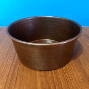 May include: A hammered copper bowl with a rounded shape and a smooth, shiny surface. The bowl is empty and sits on a wooden surface.