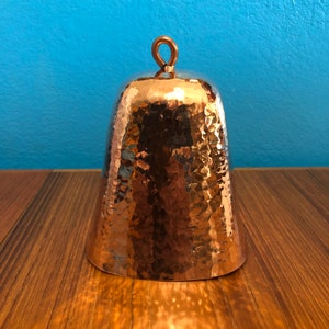 May include: A hammered copper bell with a loop handle. The bell has a rustic, aged look.