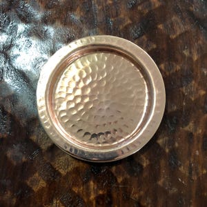 May include: A hammered copper coaster with a circular shape. The coaster has a raised rim and a textured surface with a dimpled pattern. The copper has a warm, reddish-brown hue.