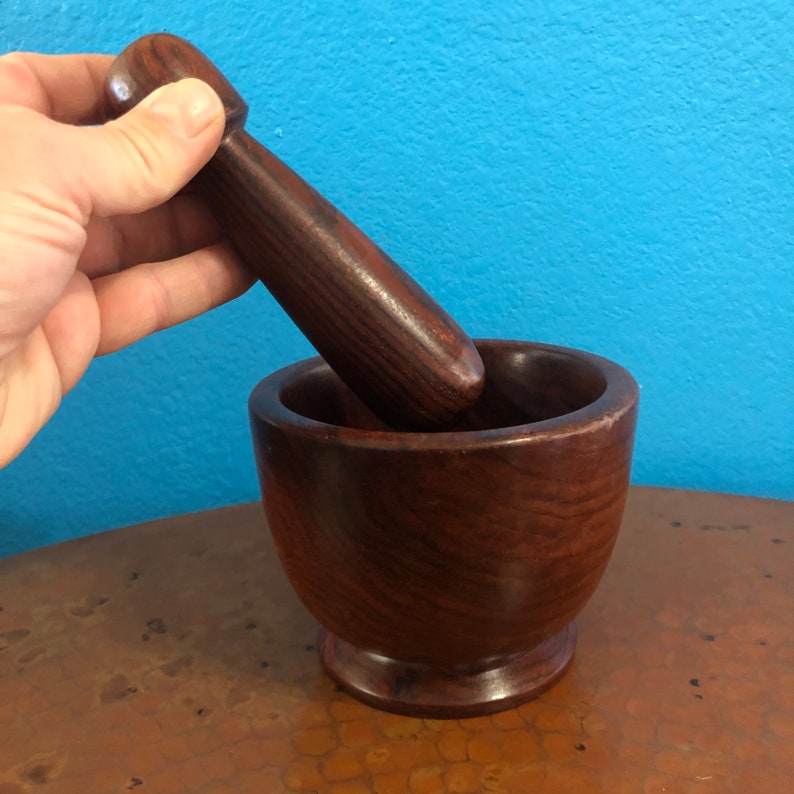 May include: A dark brown wooden mortar and pestle set. The mortar is a bowl shape with a rounded base. The pestle is cylindrical with a rounded handle. The set is made of a dark wood with visible grain.