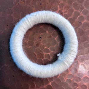 May include: A circular, white yarn wreath. The yarn is tightly wound, creating a thick, textured ring. The wreath is likely used for crafting or decoration. The background is a textured, copper-colored surface.