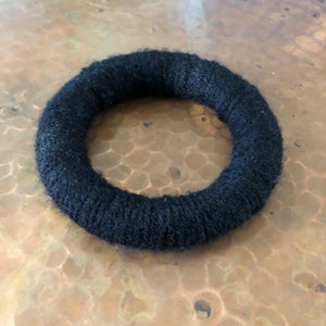May include: A black, circular hair accessory, possibly a scrunchie or hair tie, made of a soft, textured material. The hair accessory is a solid black colour and appears to be wrapped around a circular form. The item is shown against a copper-coloured surface.