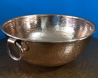 Può includere: Una grande ciotola di rame martellato con un'unica maniglia ad anello. La ciotola è lucida e ha un colore caldo metallico.