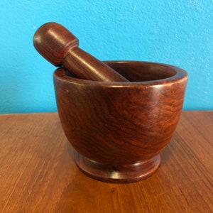 May include: A dark brown wooden mortar and pestle set. The mortar is a rounded bowl shape with a wide opening, and the pestle is cylindrical with a rounded end. The set is made of a polished wood with visible grain.
