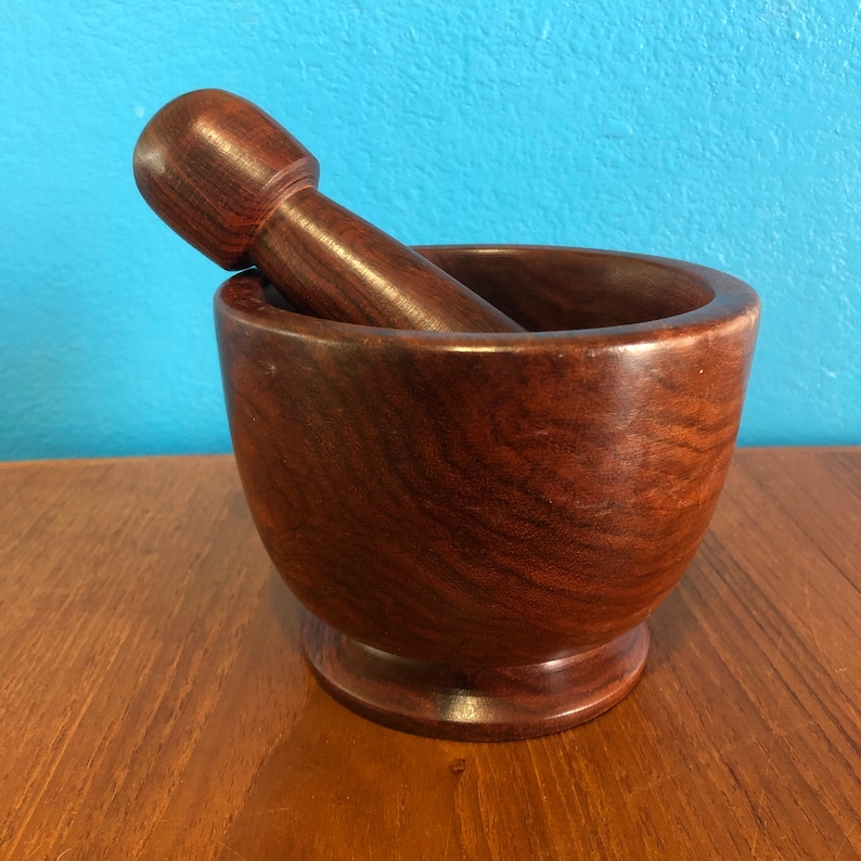 May include: A dark brown wooden mortar and pestle set. The mortar is a rounded bowl shape with a wide opening, and the pestle is cylindrical with a rounded end. The set is made of a polished wood with visible grain.