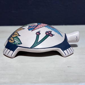 May include: A ceramic turtle figurine with a white shell decorated with colourful bird and floral designs. The turtle's head and feet are light pink, and the shell has a dark blue border. The figurine is a decorative item.
