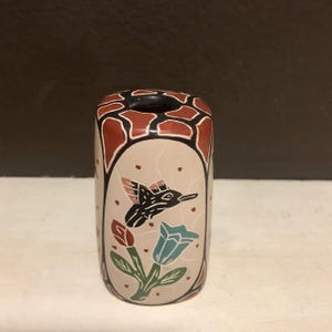 May include: A hand-painted ceramic vase with a hummingbird and floral design. The vase is cylindrical, with a beige background and a black opening. The design includes a black hummingbird, red and blue flowers, and a brown and black pattern at the top.